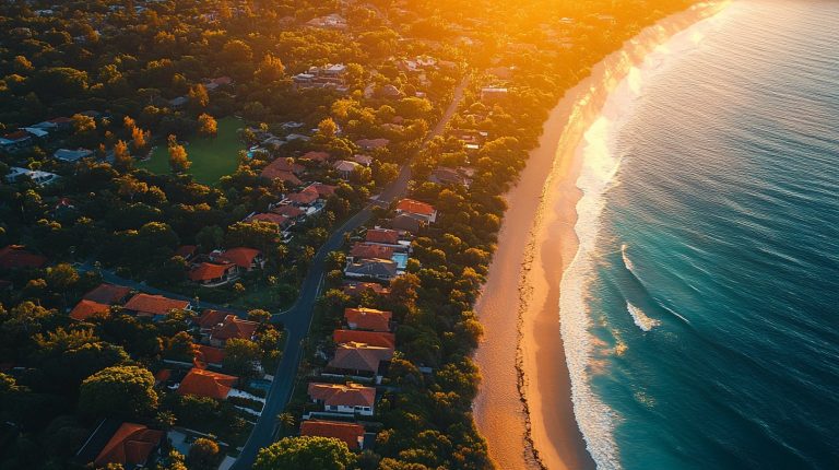 Aerial view of an Australian coastal suburb at golden hour, rooftops sweeping down to the beach